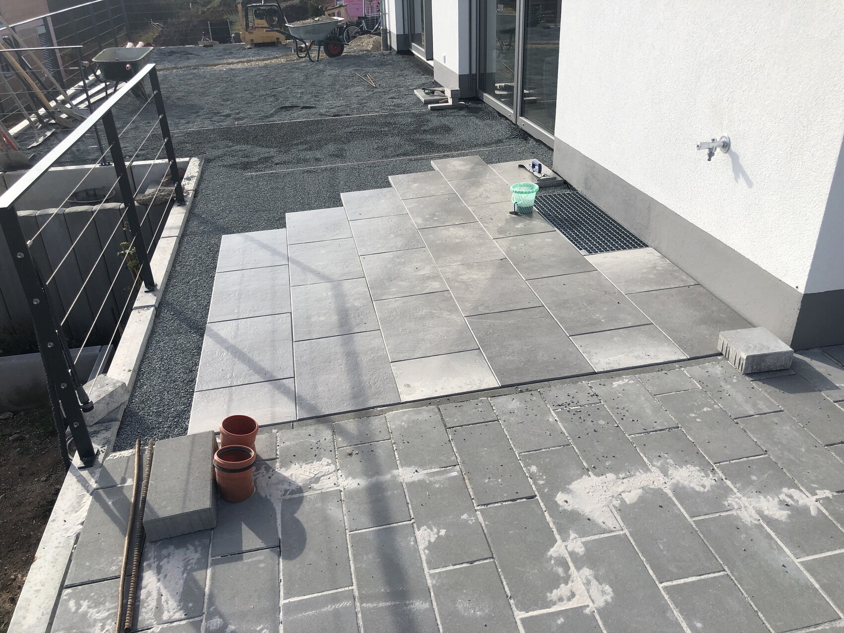 Außenbereich einer Baustelle mit grauen Pflasterplatten, Geländer links, Wasserhahn rechts.