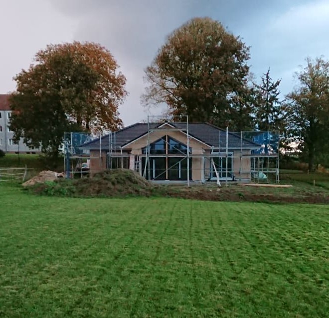 Small house under construction with scaffolding, green lawn and trees in the background.
