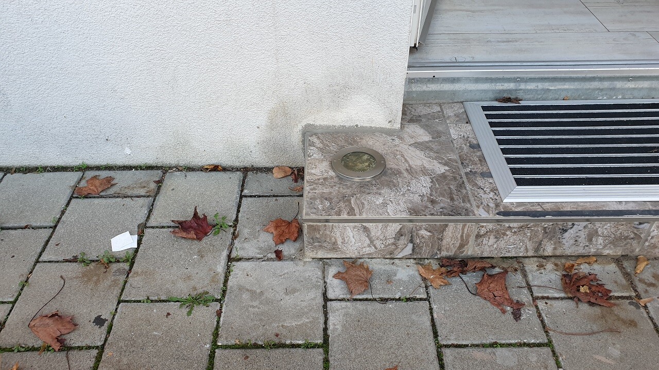 Gepflasterter Vorplatz vor dem Hauseingang mit Abflussdeckel und Herbstlaub.