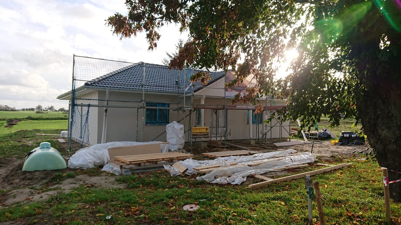 A house under construction with scaffolding, construction timber, and protective covers; tree on the right, green lawn surrounding.