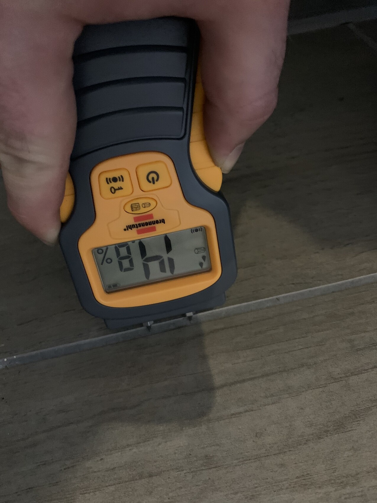 Hand holding orange moisture meter against wooden floor; display shows moisture percentage.