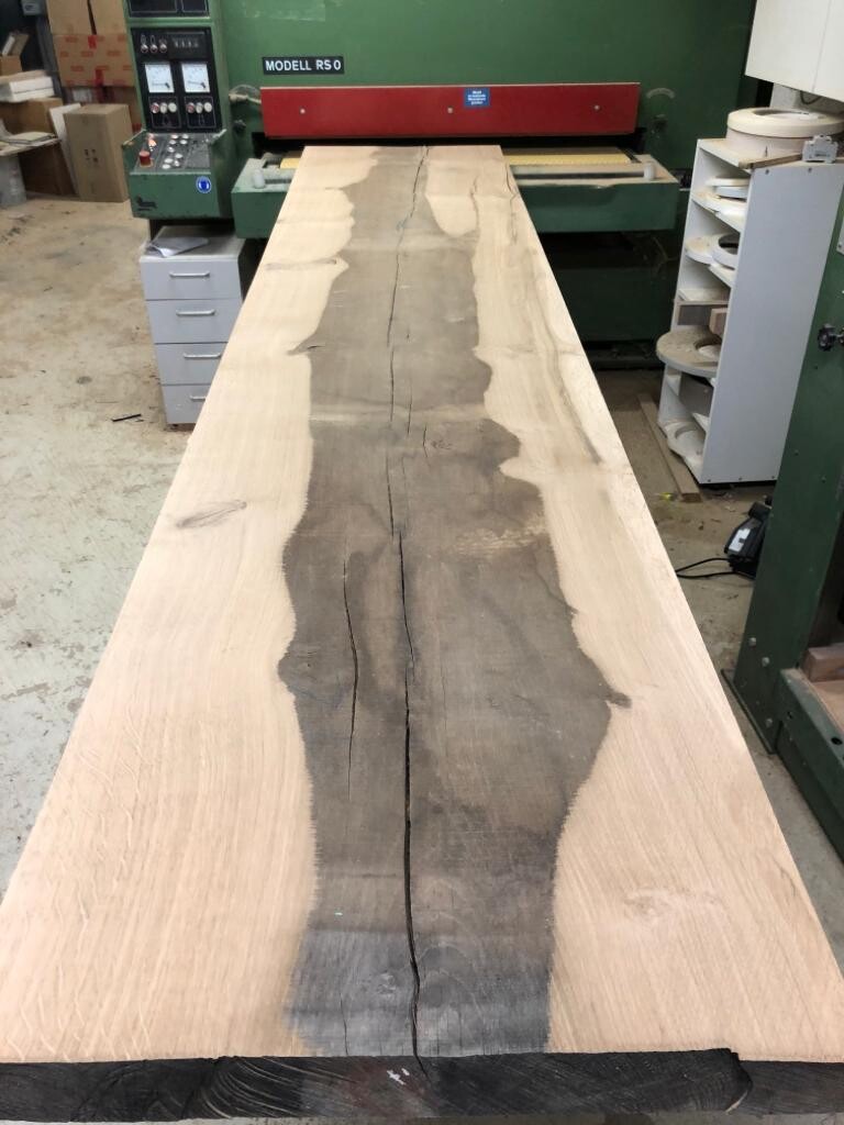Long wooden beam with a dark center and crack, in a woodworking workshop next to machines.