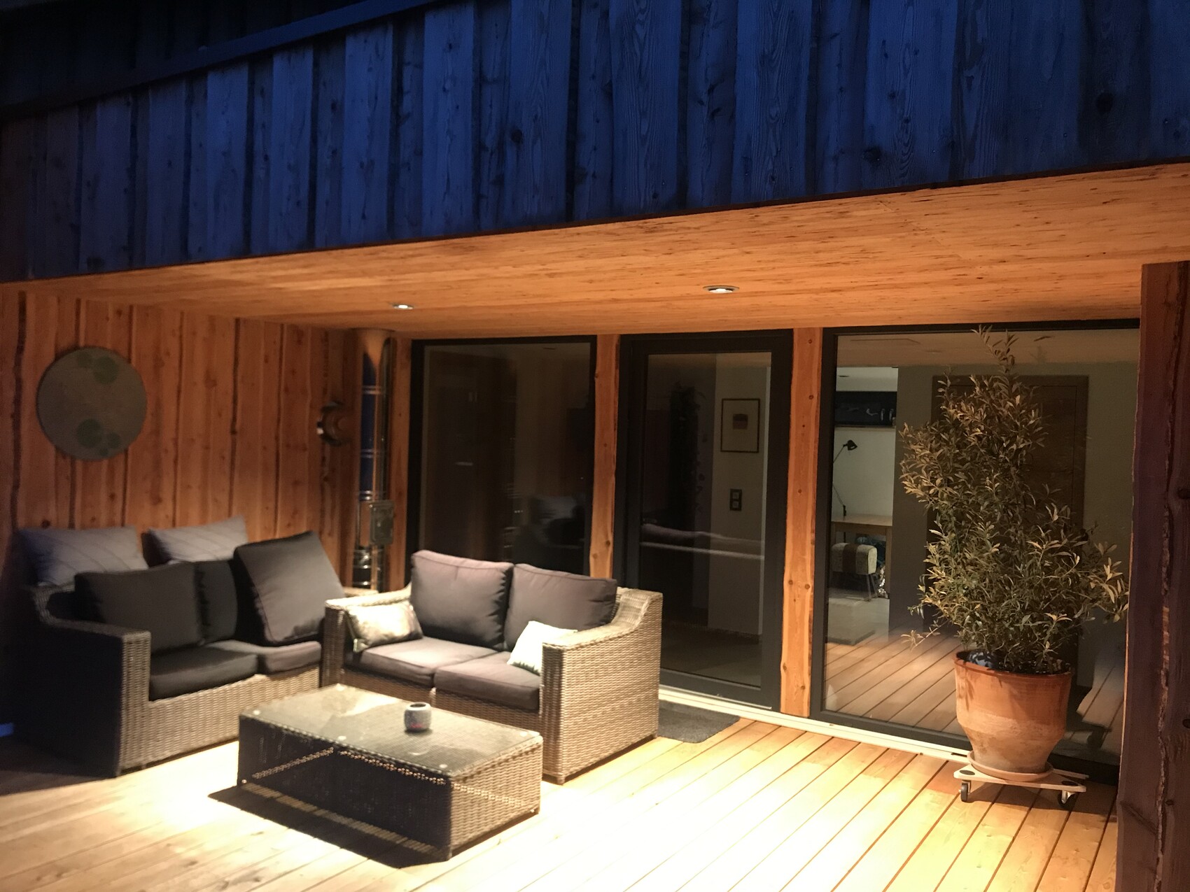 Holzveranda mit Rattan-Sofas, Glastisch und Olivenbaum vor Schiebetüren.