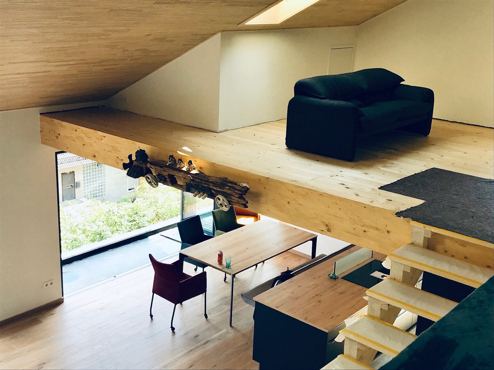 Loft with wooden floor and beams, upper lounge with black sofa, dining table below, large window.