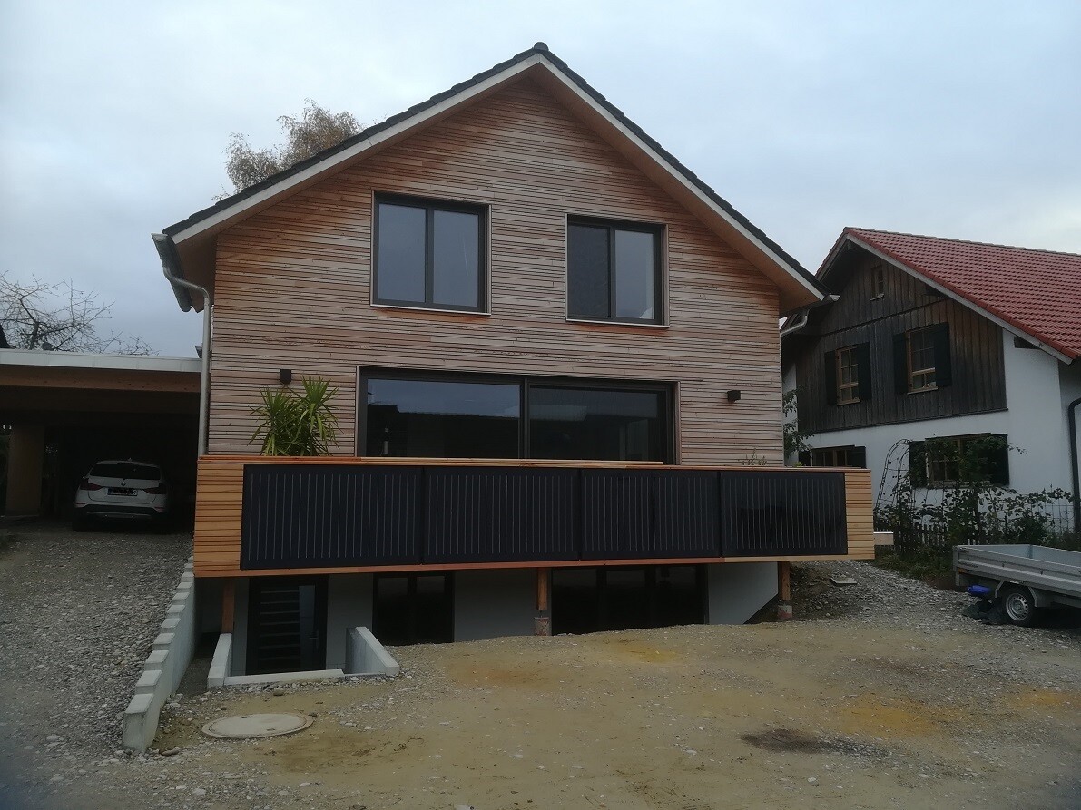 Two-story wooden house with large terrace and carport on the left.