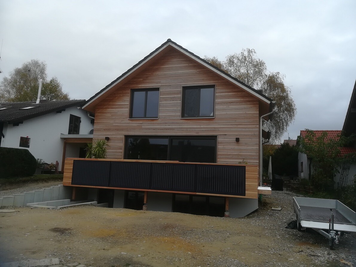 Modern wooden house with steep roof, large windows, and dark veranda on stilts.
