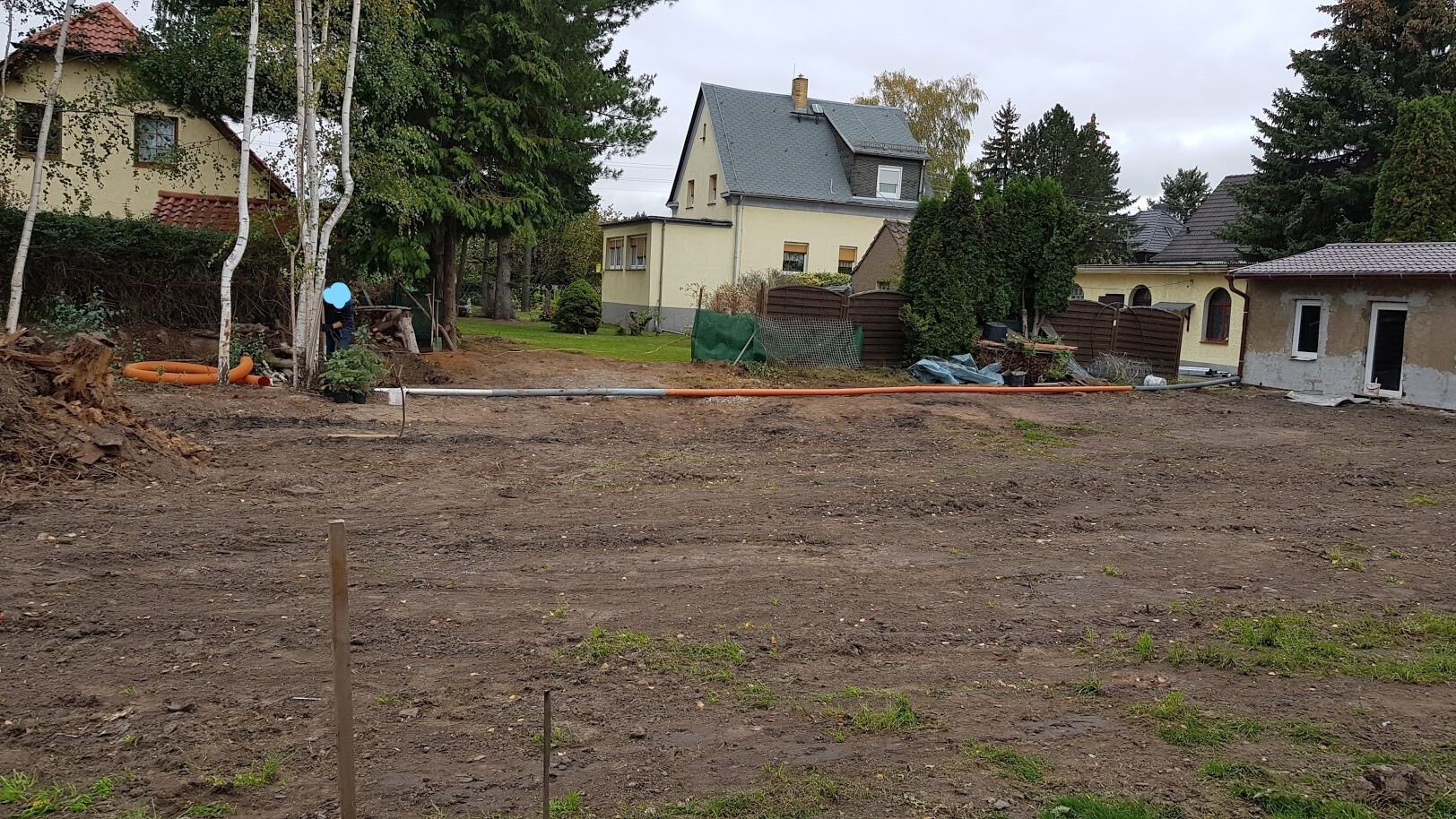 Empty construction site with earthworks, construction tools, and pipes; worker on the left, houses in the background.