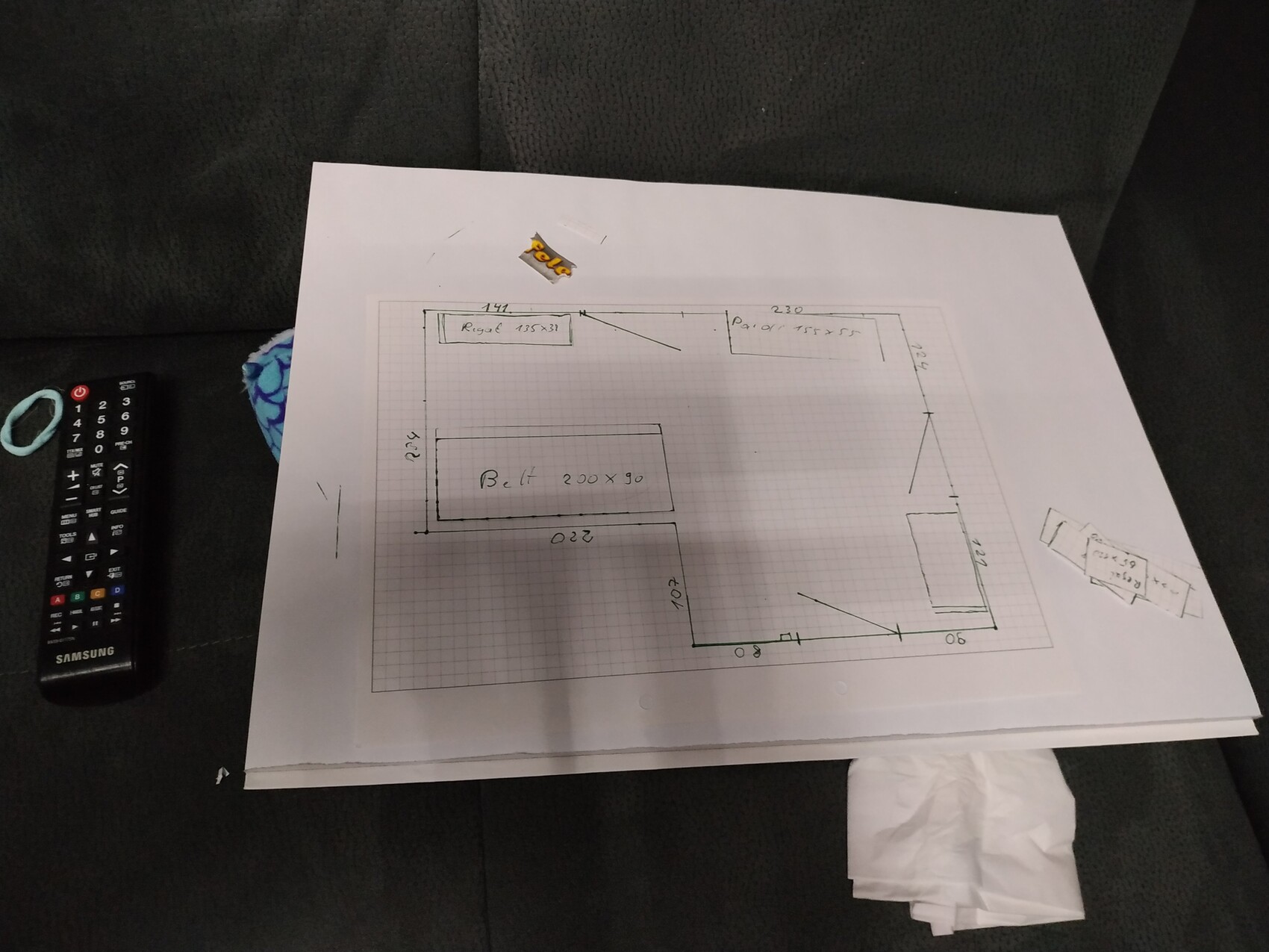 Hand-drawn floor plan sketch with room layout on paper, next to a remote control on a dark sofa