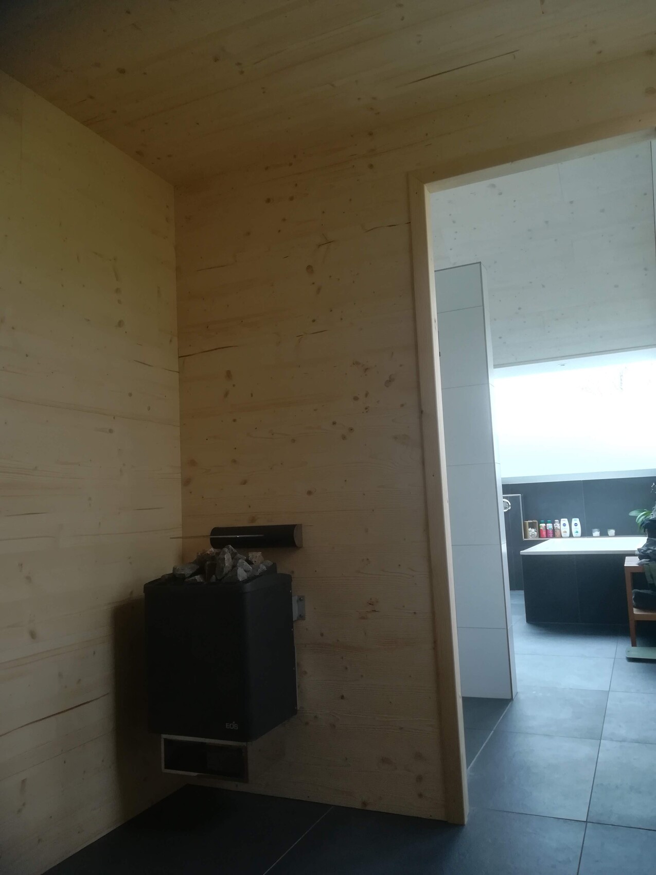 Wood-paneled room with sauna heater and stone top; view into adjoining kitchen corner with tiles.