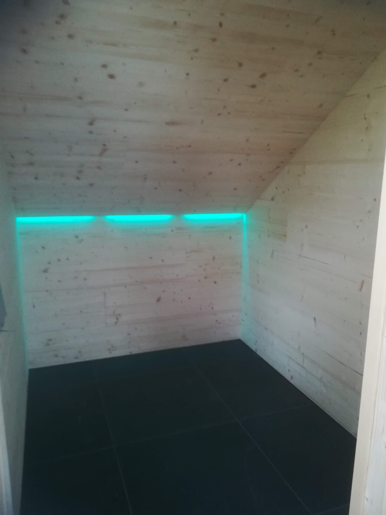 Small room with wood walls and ceiling, dark floor; turquoise LED light strip on the wall.