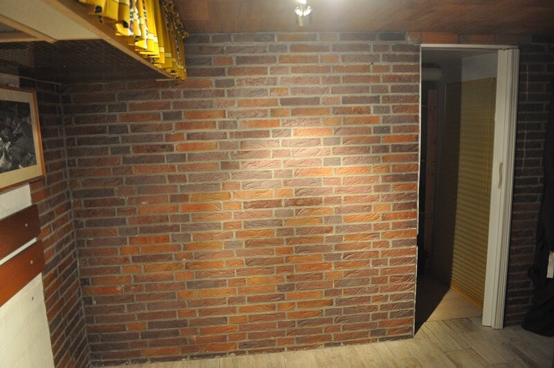 Interior with brick wall, wooden floor; door on the right to adjacent room, yellow shelf on the left.