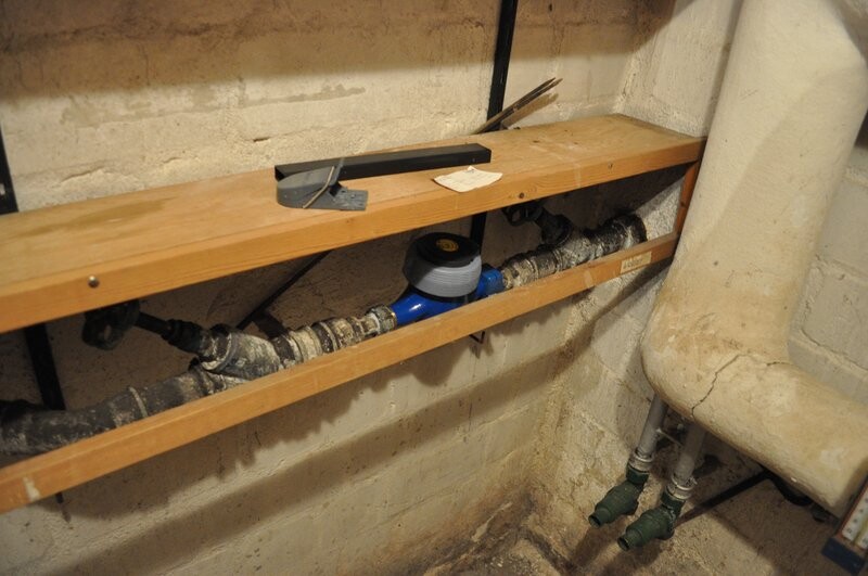 Basement room with wooden shelf above pipes, blue measuring device/shut-off valve, and insulated pipes.