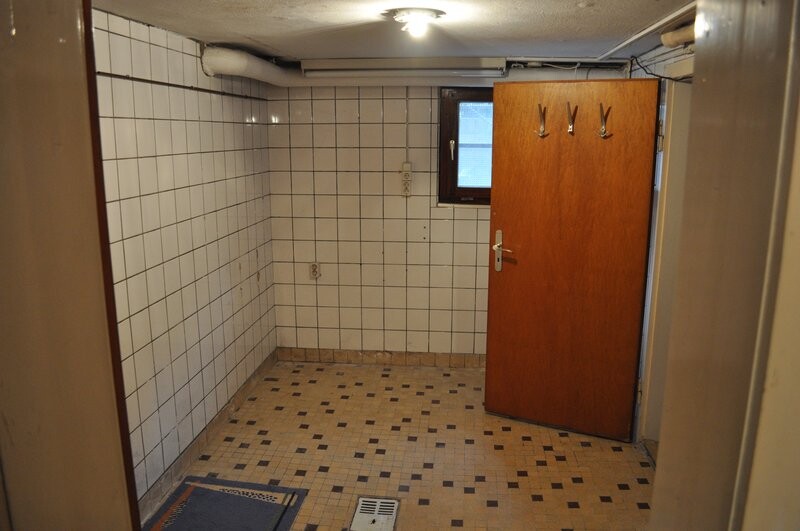 Basement room with white wall tiles, wooden door with hook, small window, and floor tiles.