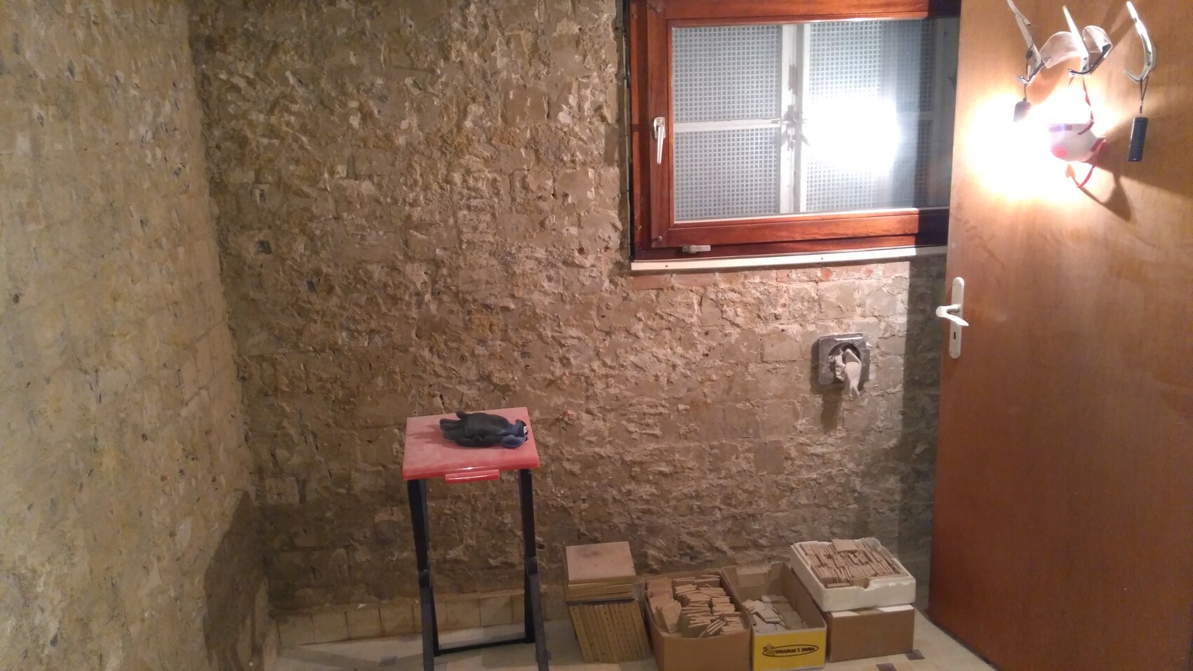 Basement room with rough stone wall, window, wooden frame; red table with gloves, boxes with tiles.