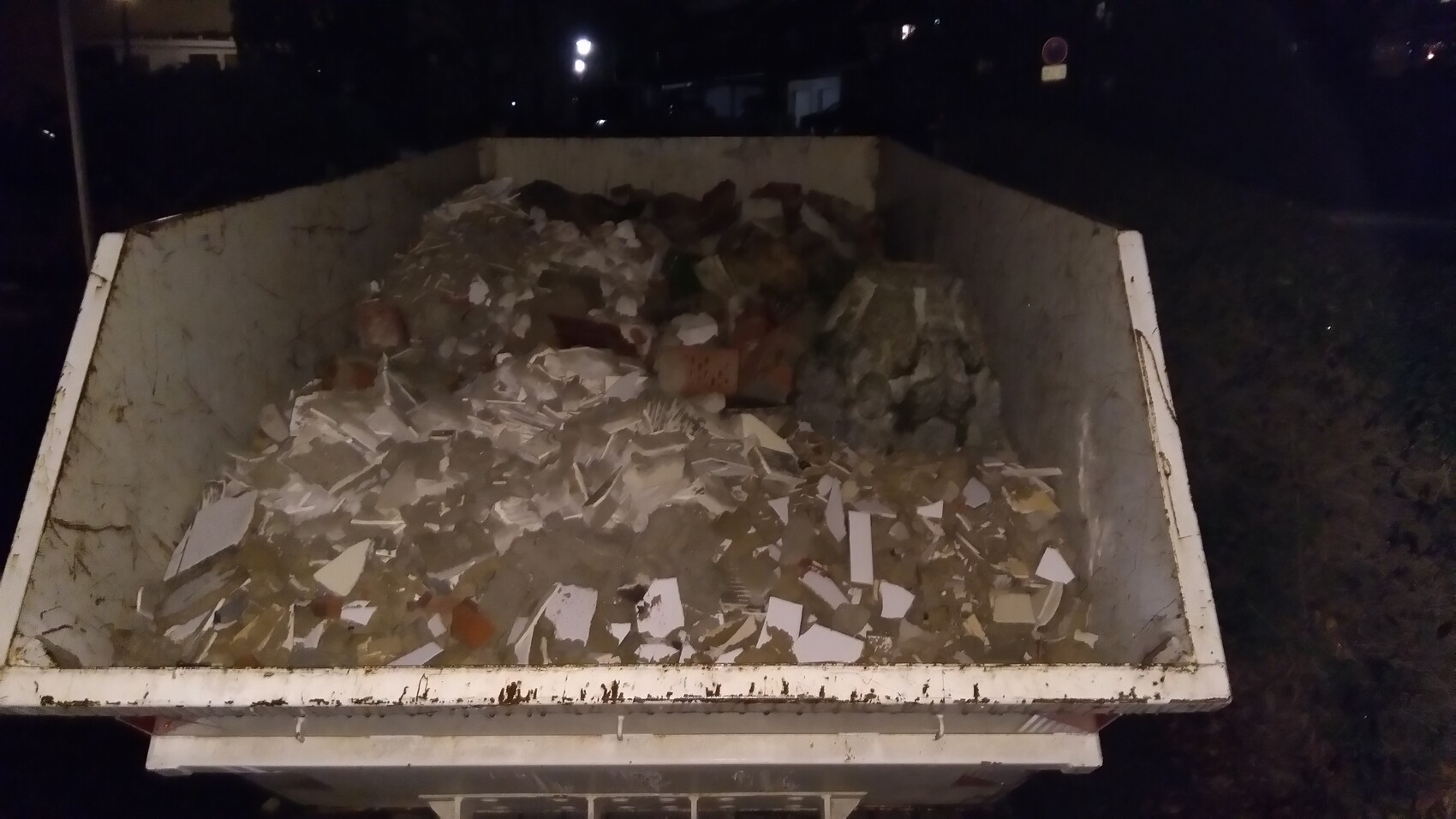 Night shot of a full waste container with construction debris, tiles, and paper.