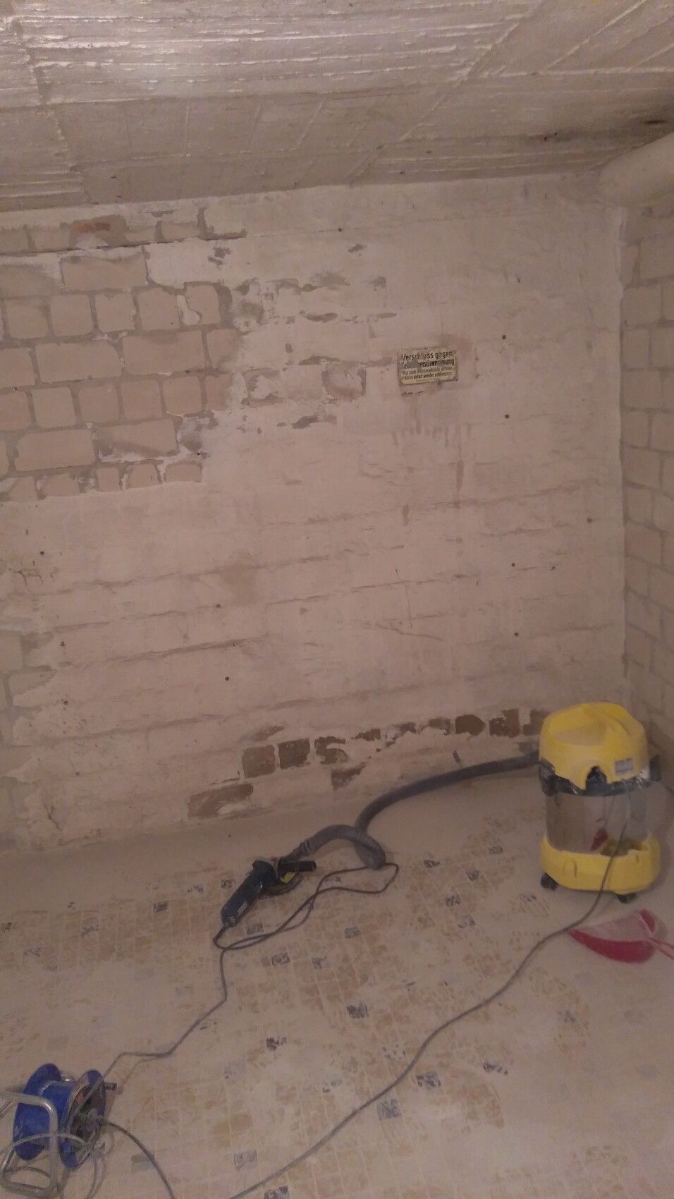 Basement room with white plastered brick wall, dust, and a yellow wet-dry vacuum cleaner with cables.