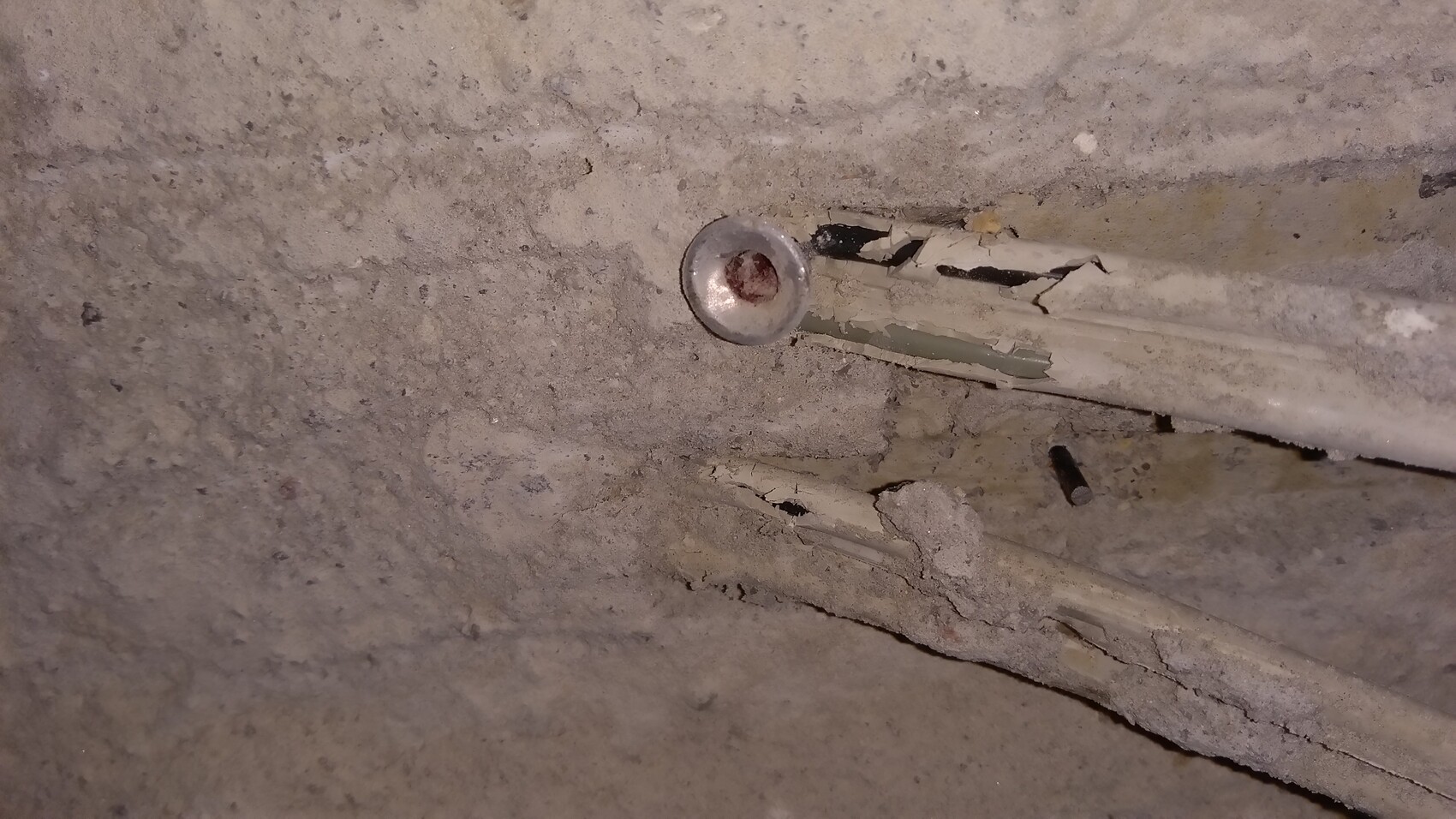 Round metal cap in dusty concrete wall, with crumbling conduit and cracks nearby.