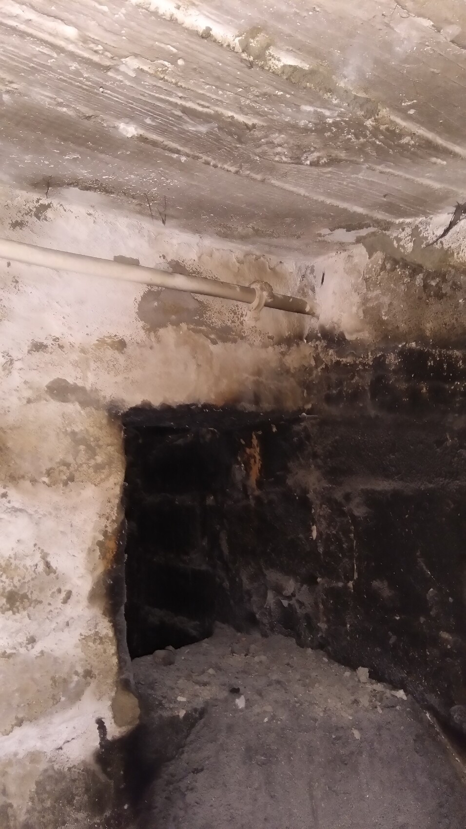 Basement room with rough wall, pipe on the ceiling, dark opening in the wall, dusty floor.