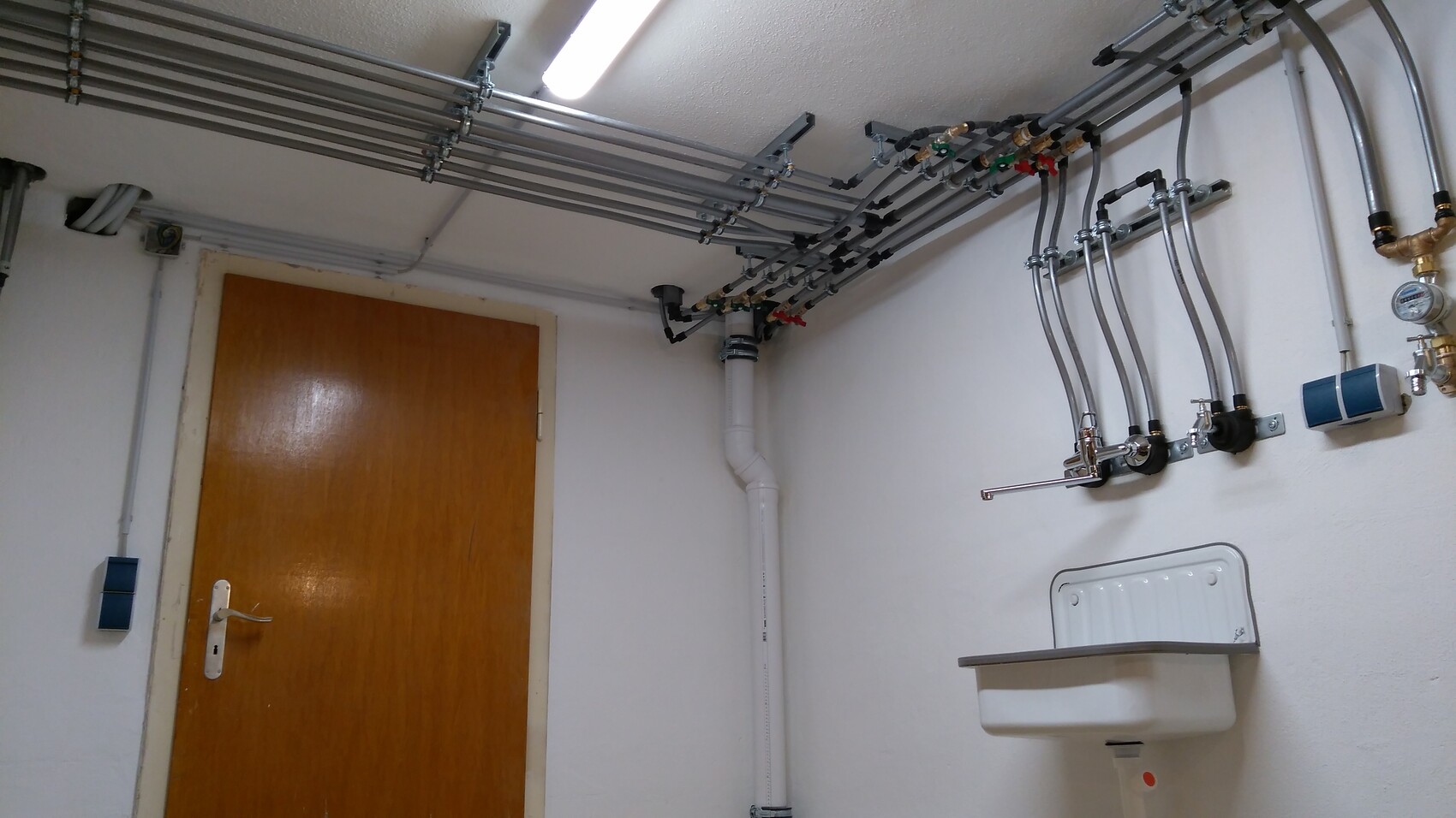 Utility room with many pipes on the ceiling, valves, measuring device, sink, and door.