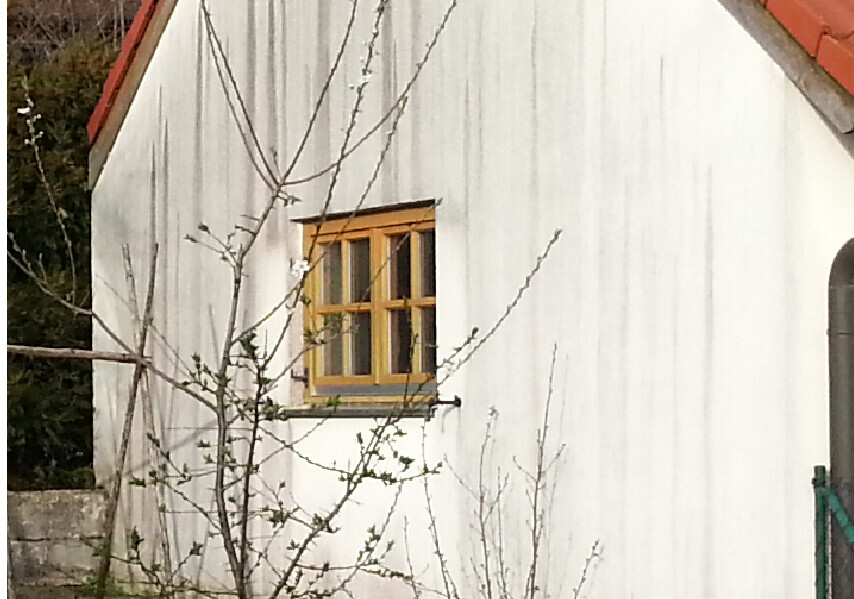 Weiße Hauswand mit kleinem Holzfenster im orange-gelben Rahmen; Baumzweige davor.