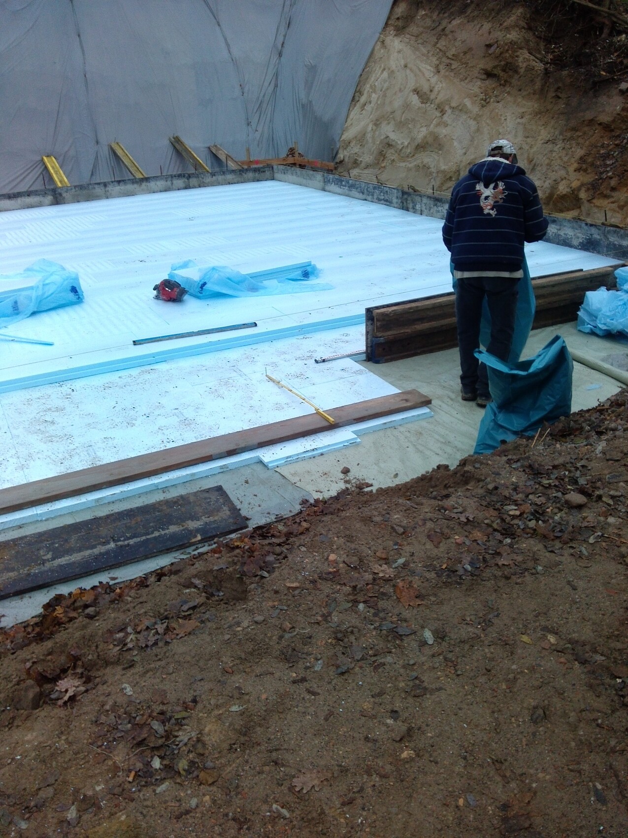 Construction worker installs white insulation boards in excavation; surrounding soil and tools visible.
