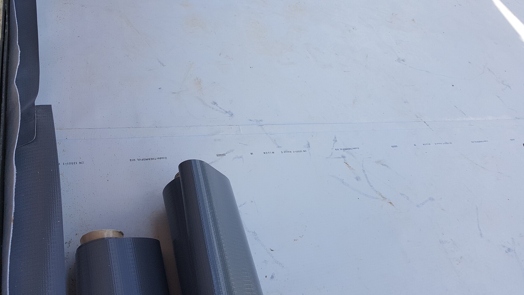 Several dark rolls of roofing membrane materials lying on a light gray surface, left side.