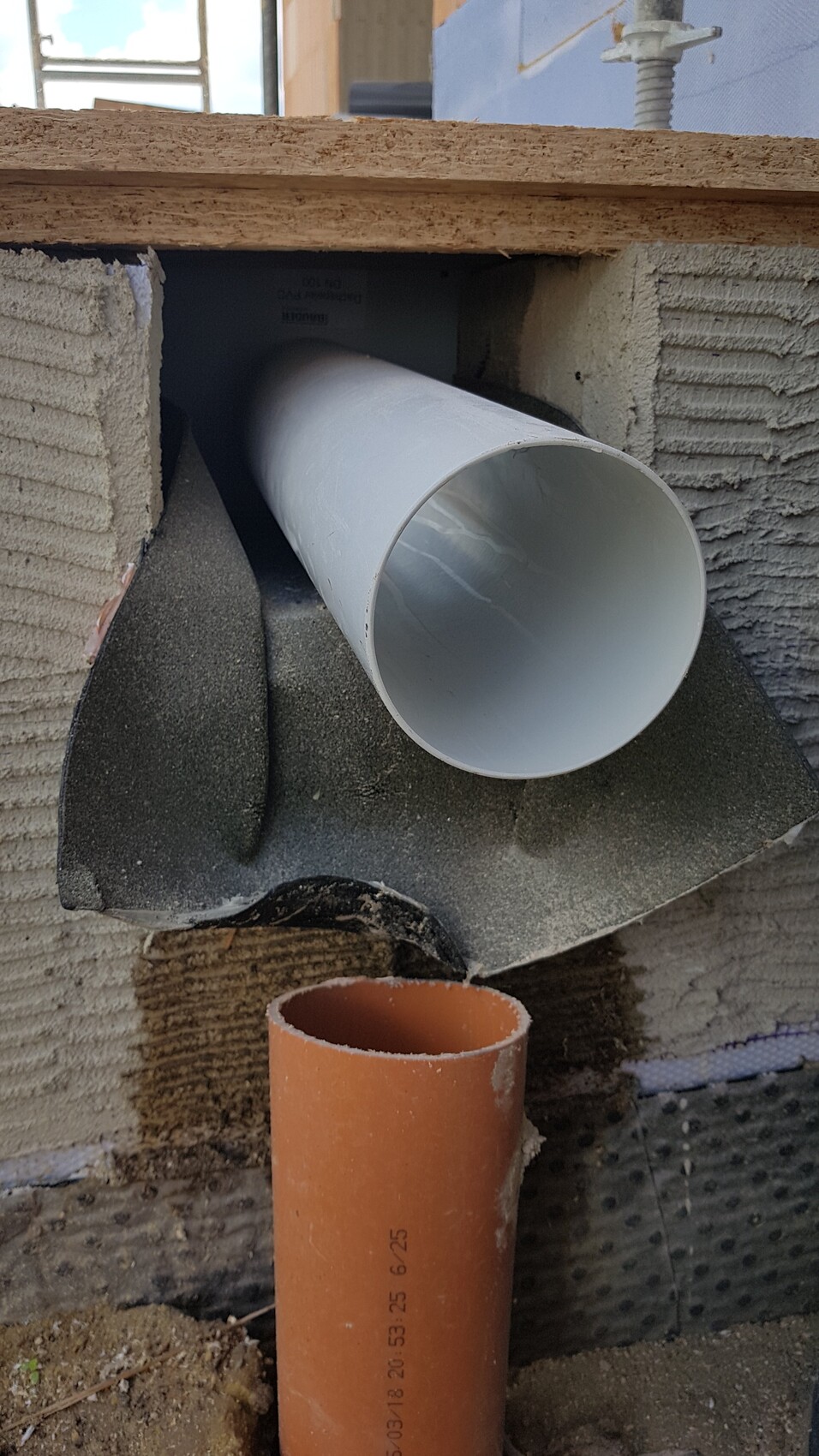 White PVC pipe running horizontally through a wall opening; orange pipe underneath.