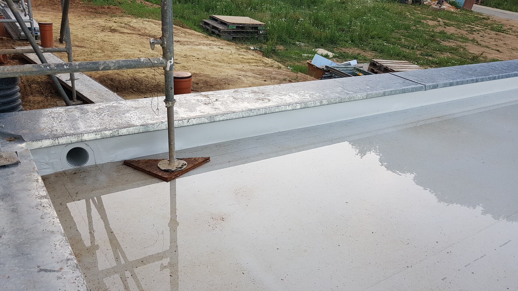 Construction site: wet concrete surface with drainage opening, support pipe on wooden panel, scaffolding in background.