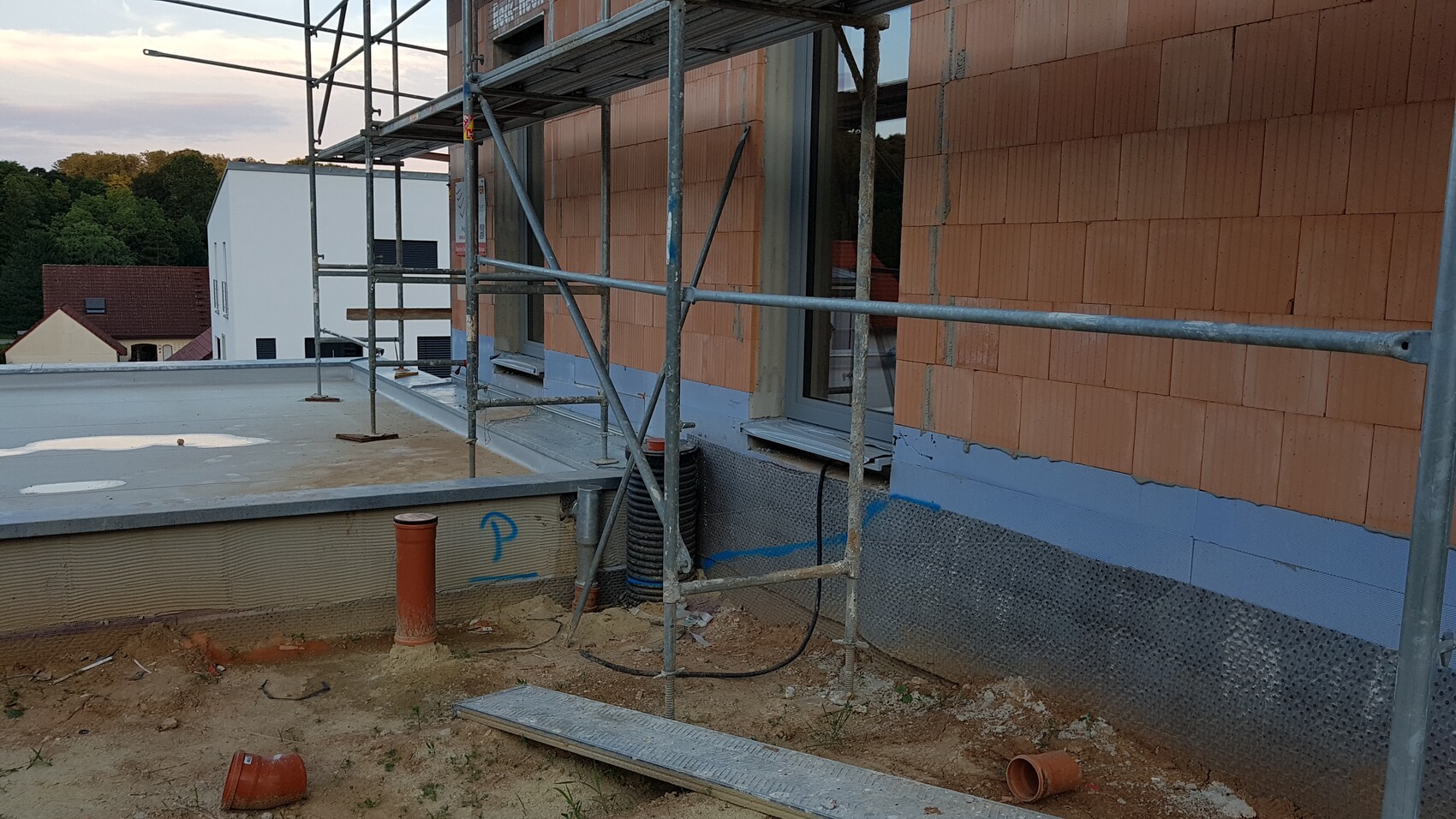 Building scaffolding in front of orange brick wall of a building, windows, earthworks and construction debris visible.