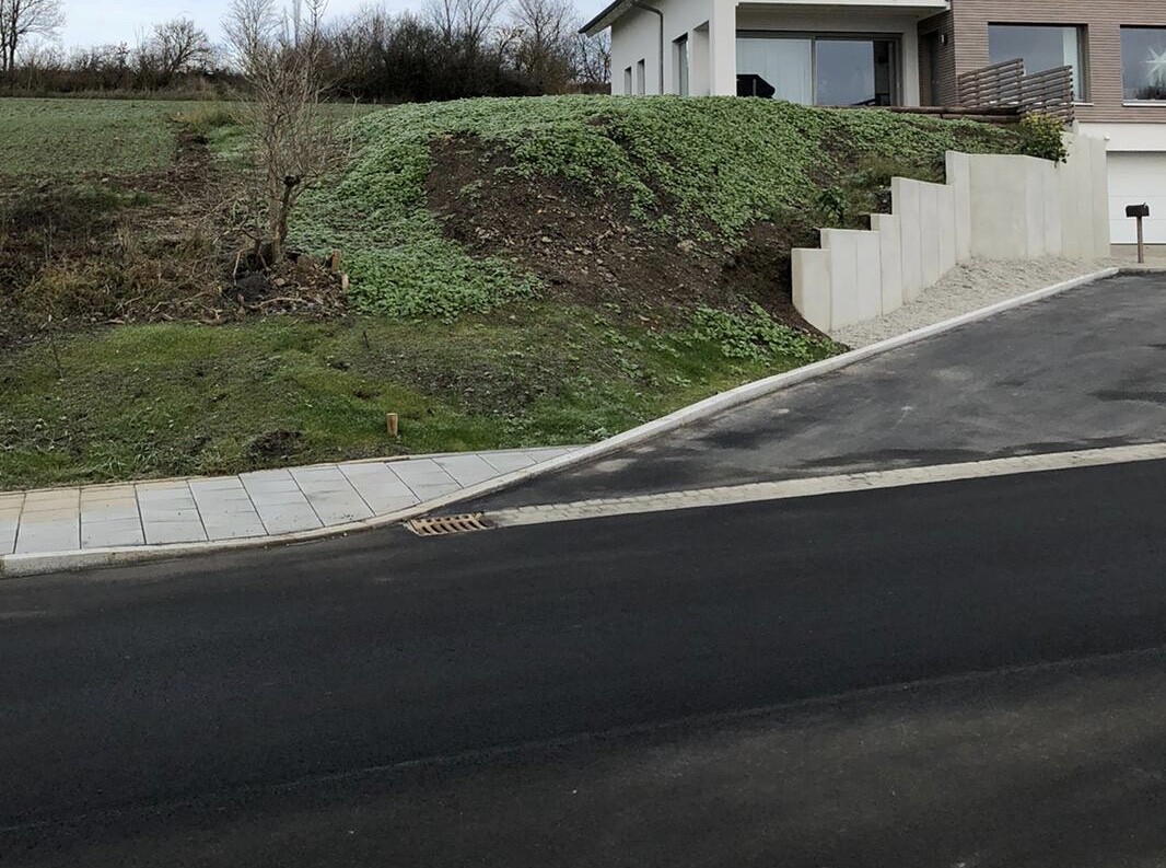 Slope with greenery next to modern house; walkway, street and retaining wall made of concrete.