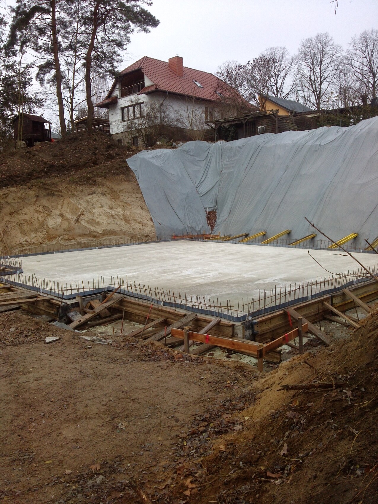 Baustelle mit frisch gegossener Bodenplatte, Stahlbewehrung, Holzformwerk, Haus im Hintergrund.
