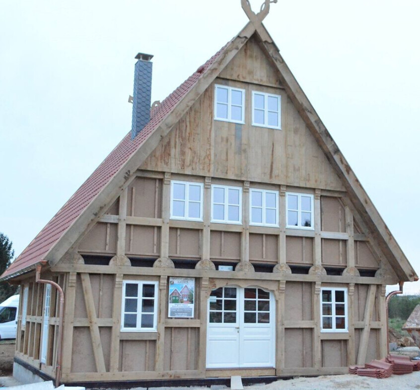 Timber-framed house under construction with red gable roof, white windows and doors, visible wooden beams