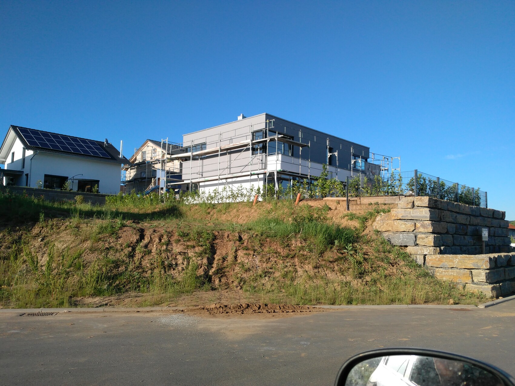 Neubau auf Hügel mit Gerüst, daneben kleines Haus mit Solarmodulen; Steinmauer rechts; blauer Himmel.