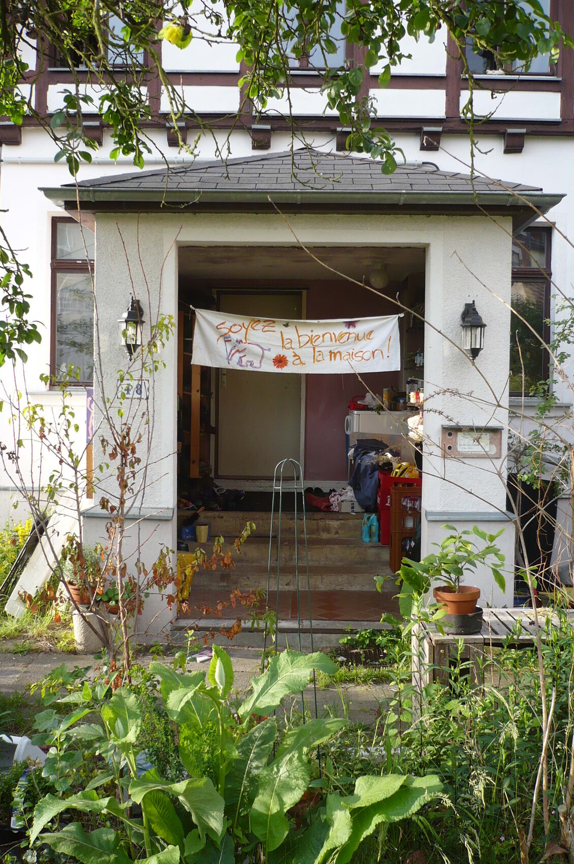 Open front door with banner 'soyez la bienvenue à la maison' in front of garden plants.