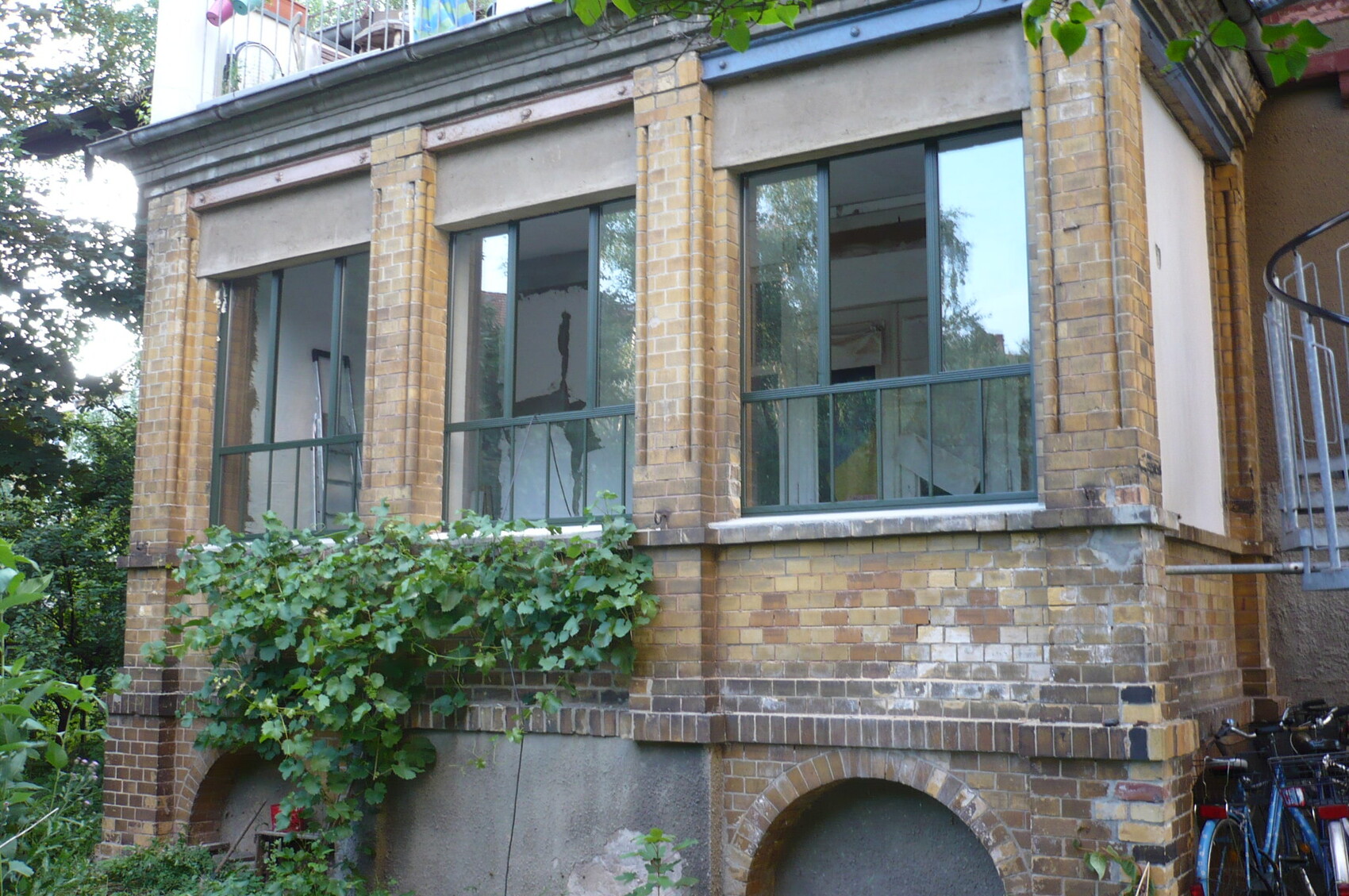 Außenansicht eines gelb-braunen Backstein-Gebäudes mit drei Fensterfronten, Rankpflanzen und Fahrrädern rechts.