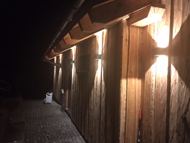 Nächtliche Holzfassade entlang einer kopfsteinernen Gasse; warme Wandbeleuchtung, linke Tasche sichtbar