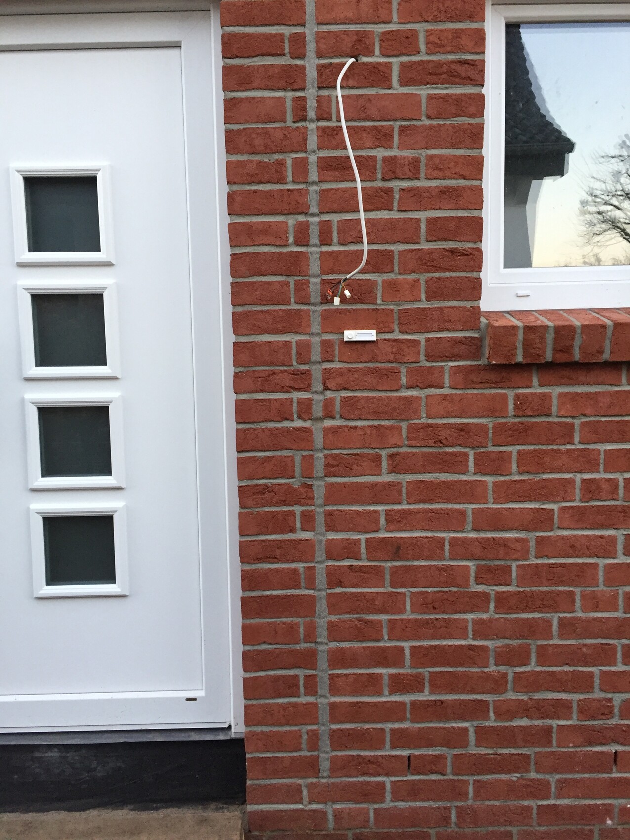 Außenansicht eines roten Ziegelhauses mit weißer Tür und Fenster, lose Kabel an der Wand.