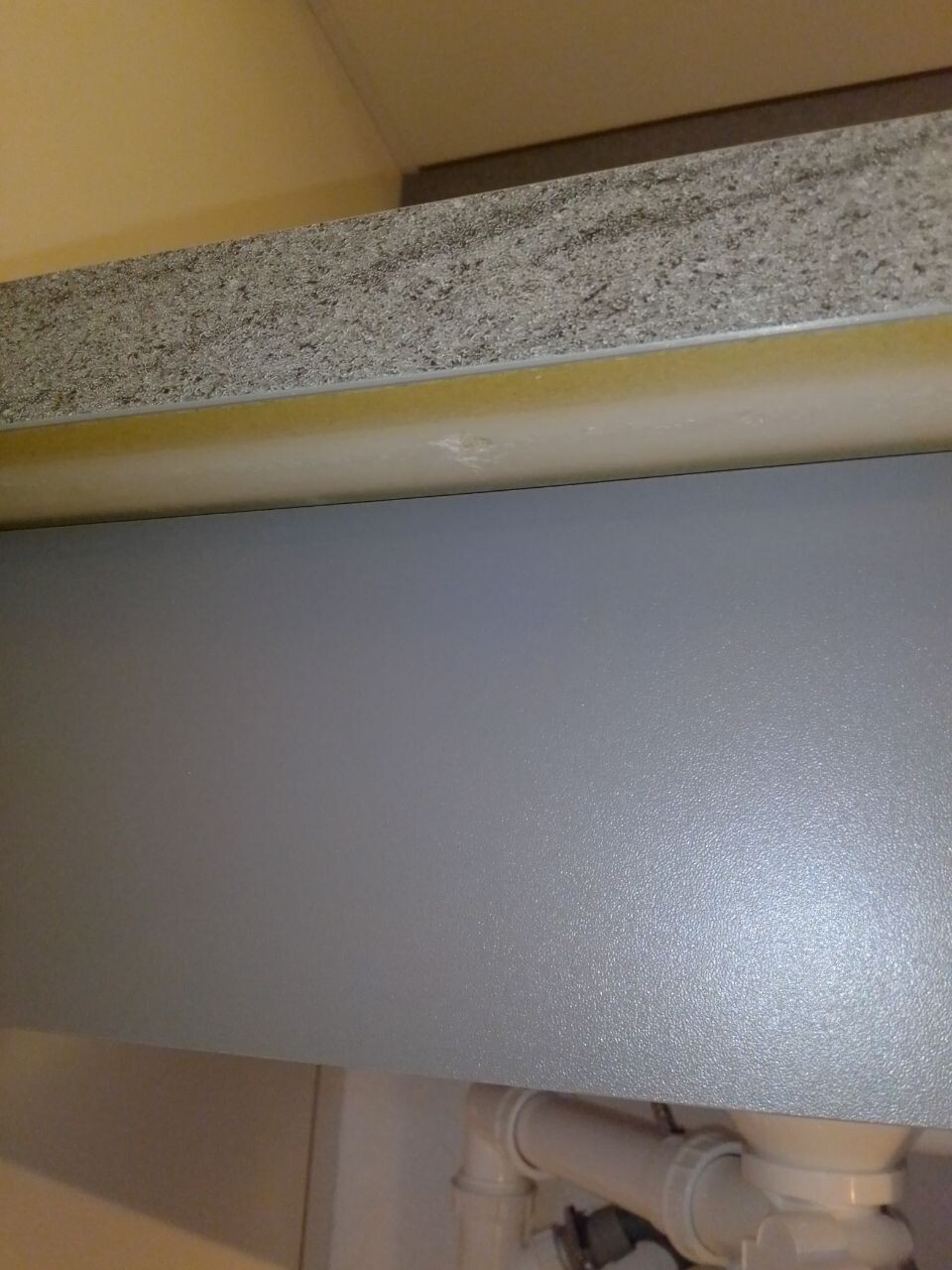 Unter dem Spuelenschrank sichtbare Rohrleitungen und Wasserinstallation hinter grauer Arbeitsplatte