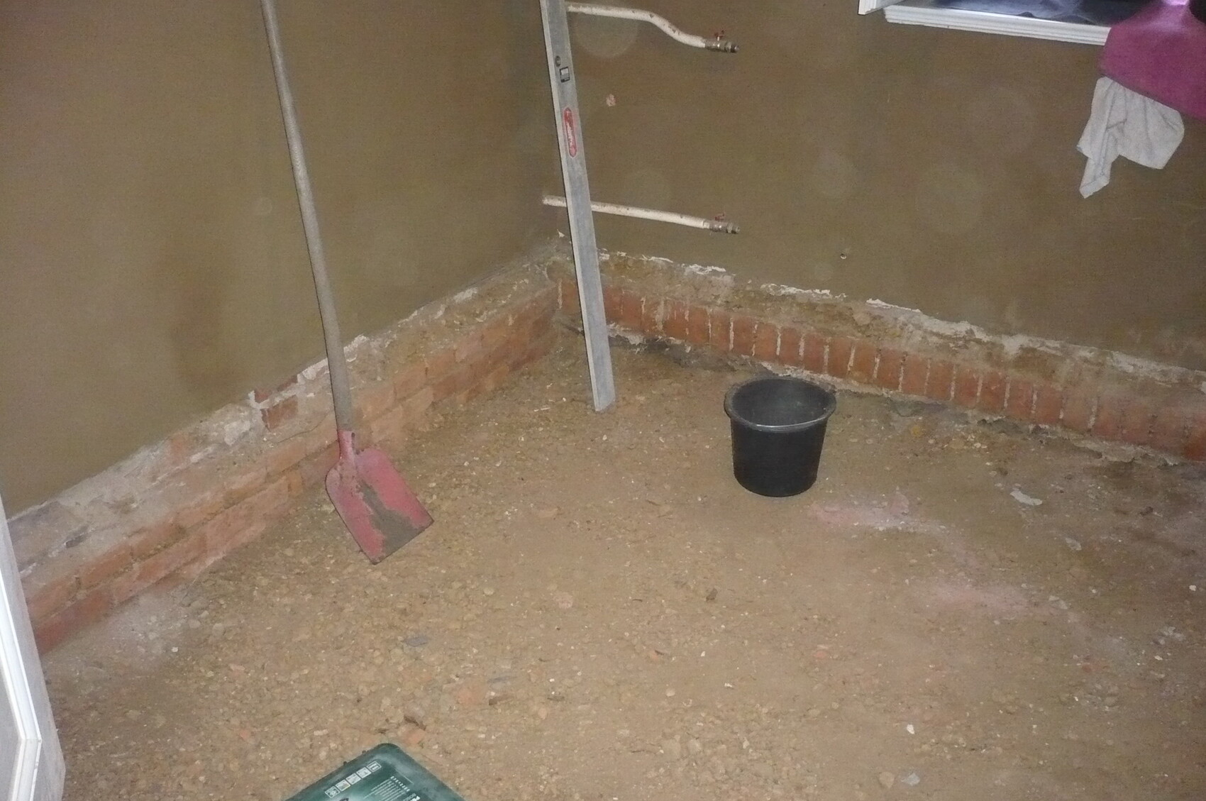 Cellar room with gravel floor, red shovel, black bucket, and ladder in front of rough brick wall.
