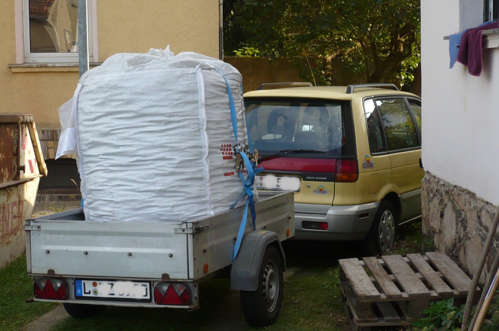 Beige van with trailer loaded with a large white sack, pallets next to it.