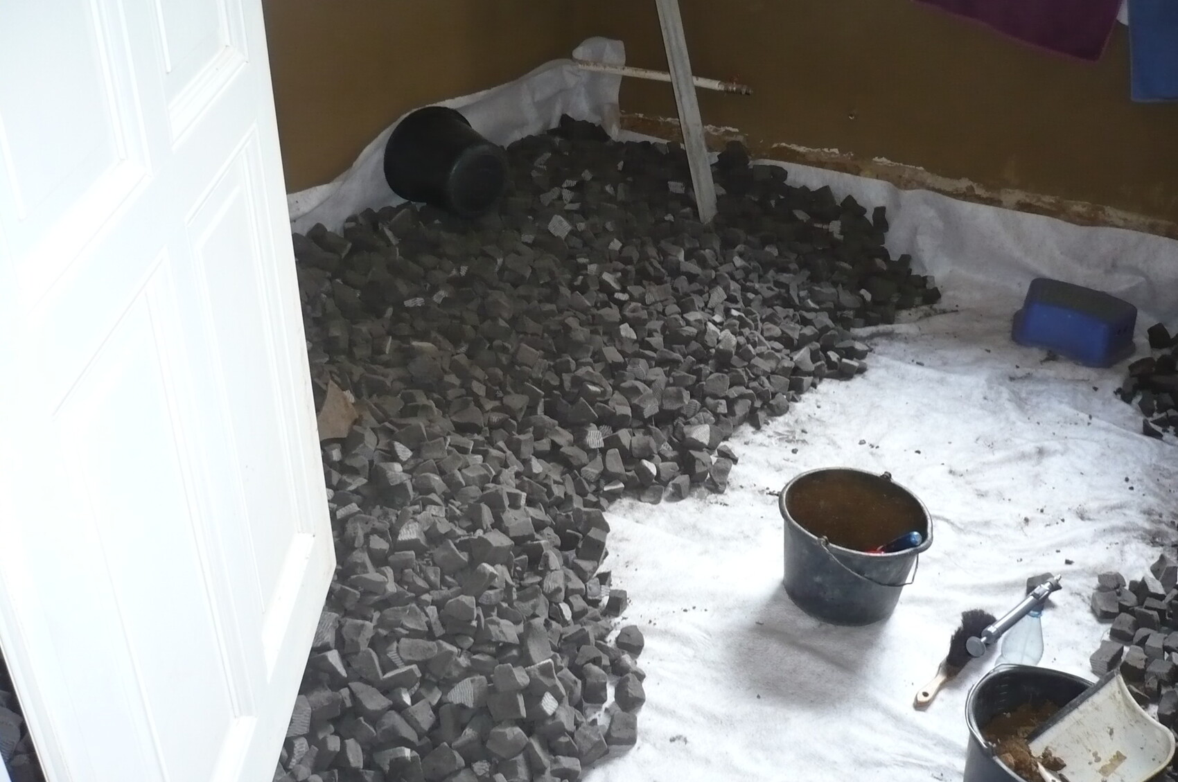 Cellar room with white floor membrane, large pile of black stones, bucket, and tools.