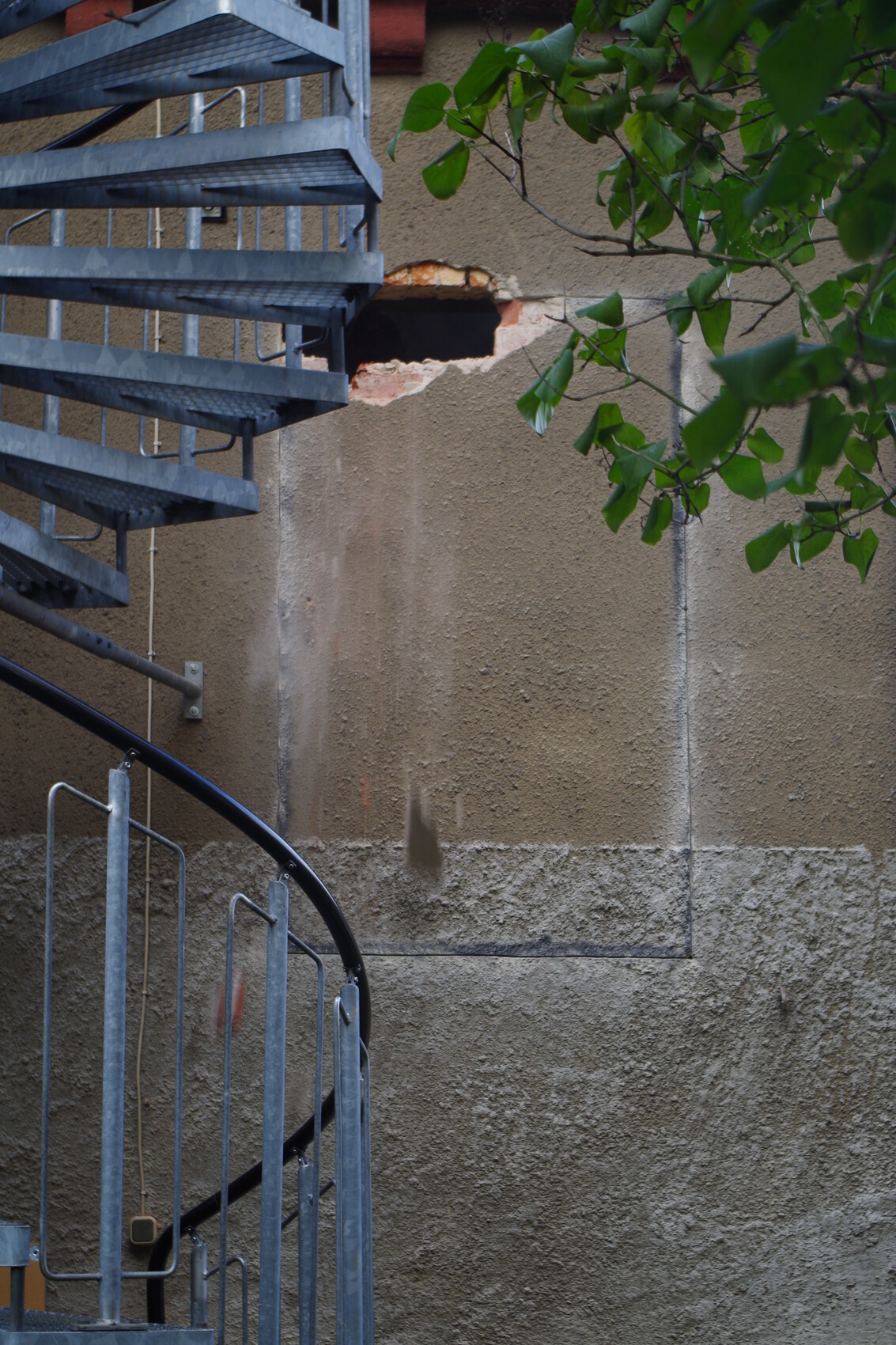 Exterior wall with damage to the plaster surface, next to a blue metal staircase and green leaves.