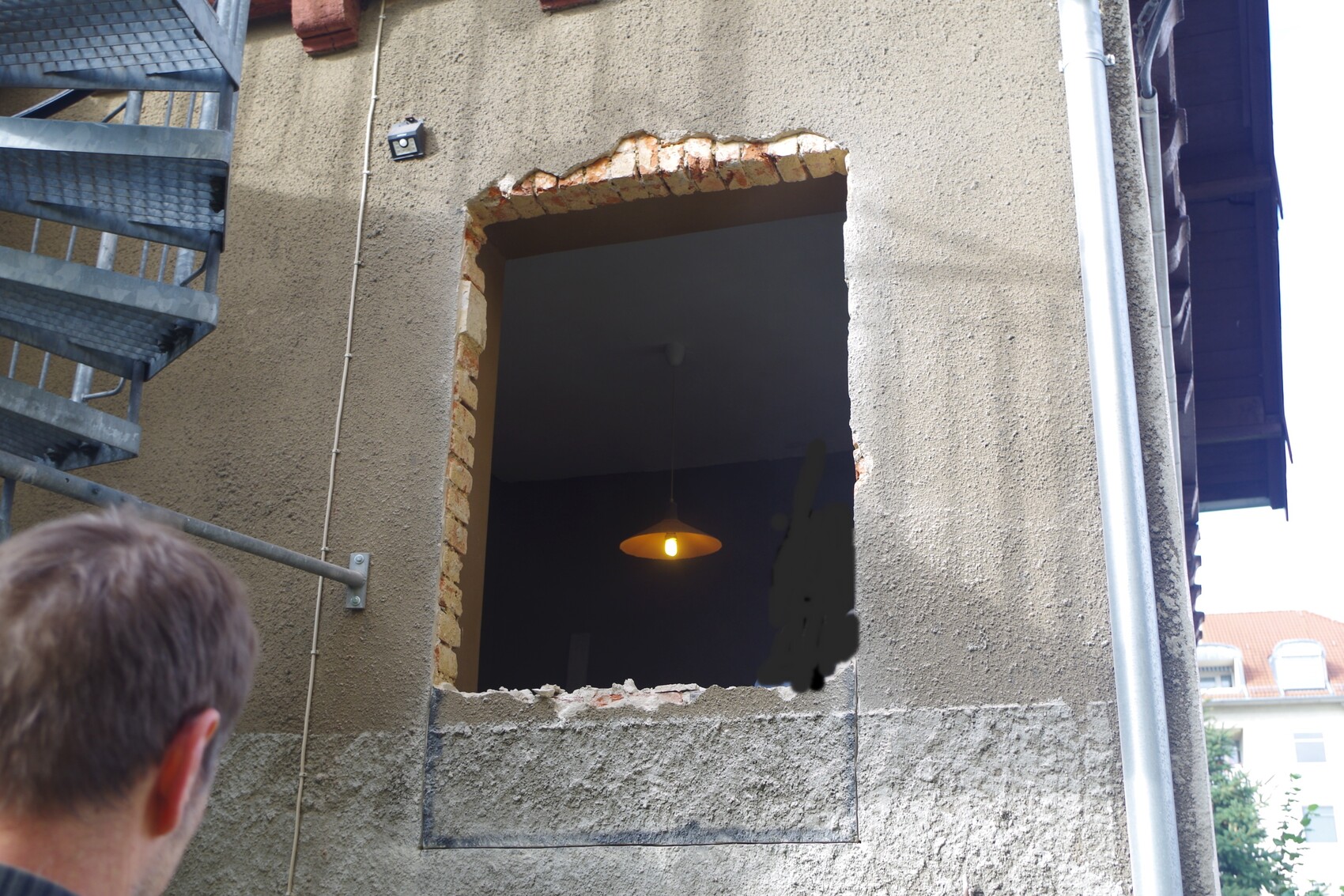 Exterior wall with open window opening made of rubble stone; dark interior, yellow pendant light; person from behind.