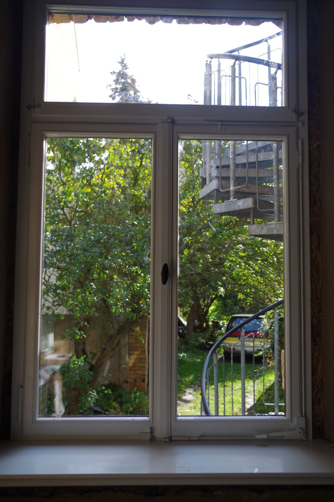 View through window to garden with trees, metal external staircase on the right, car in the background.