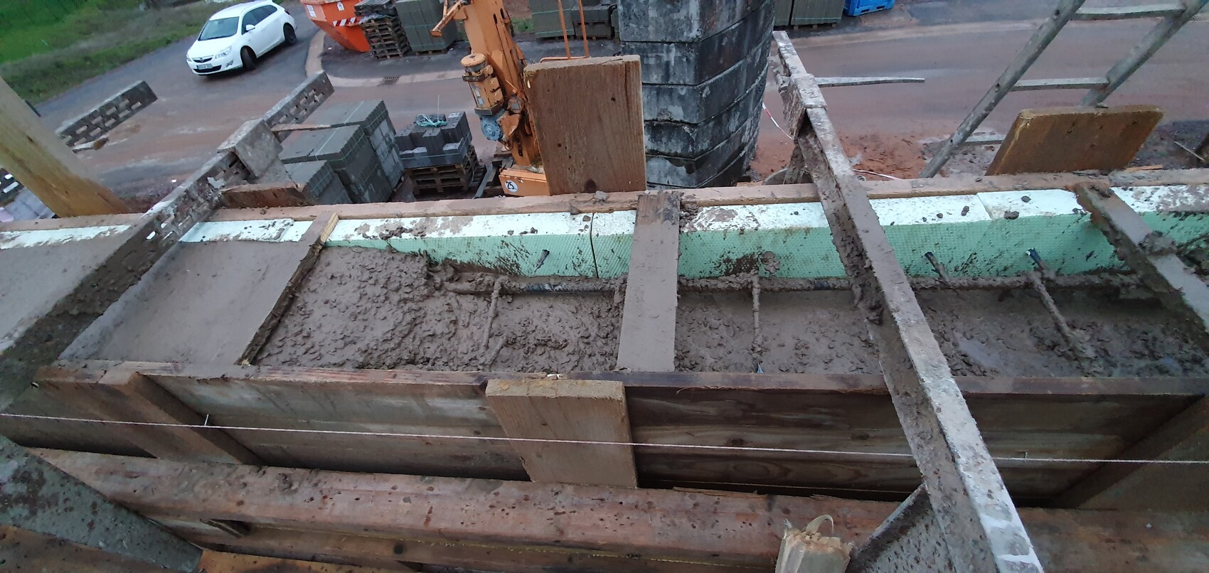 Baustelle: Holzschalung, schlammiger Beton, Bewehrung, orange Kran, Paletten.