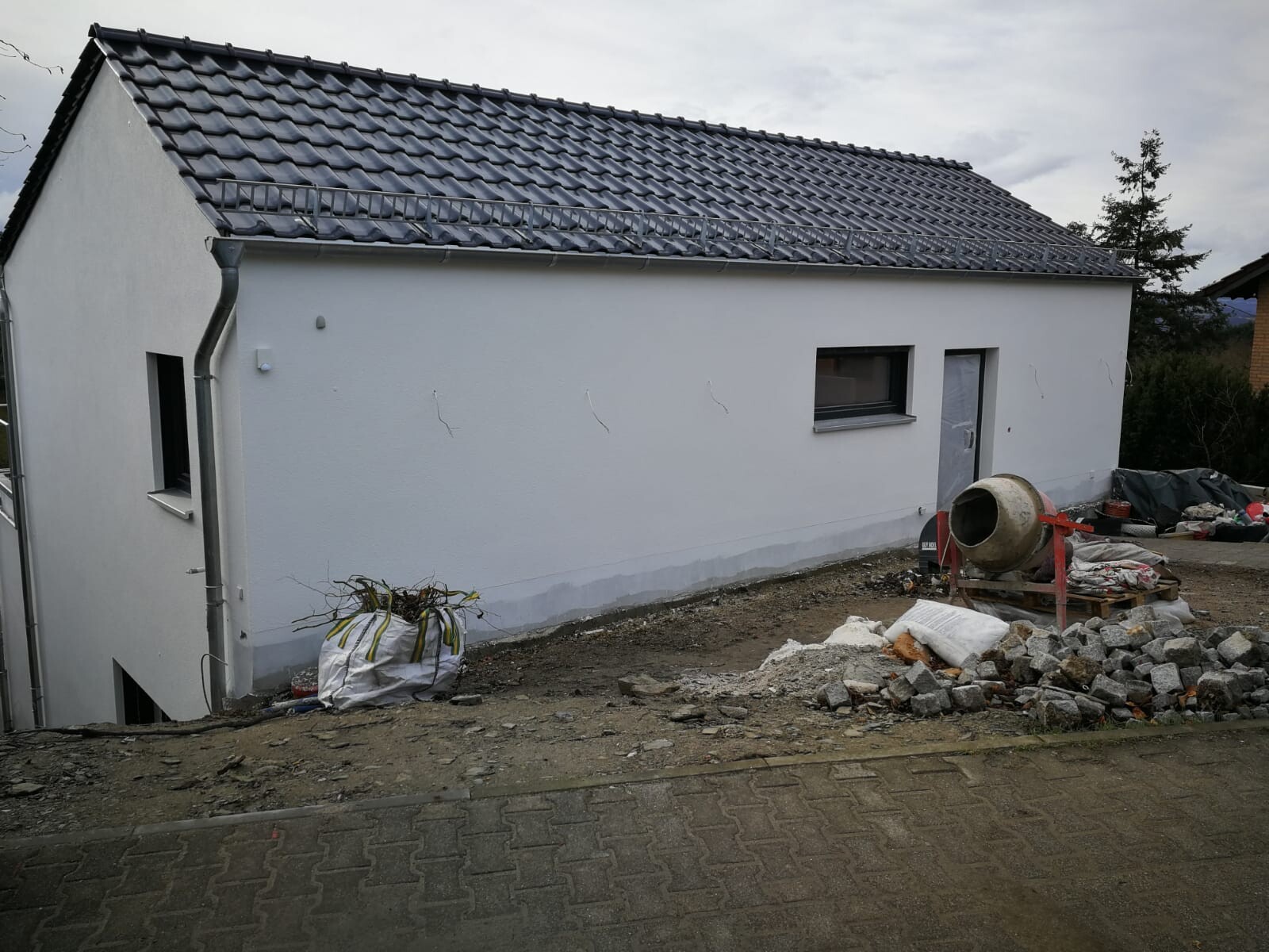 Small white house with dark tiled roof at construction site, building materials and cement mixer in yard.