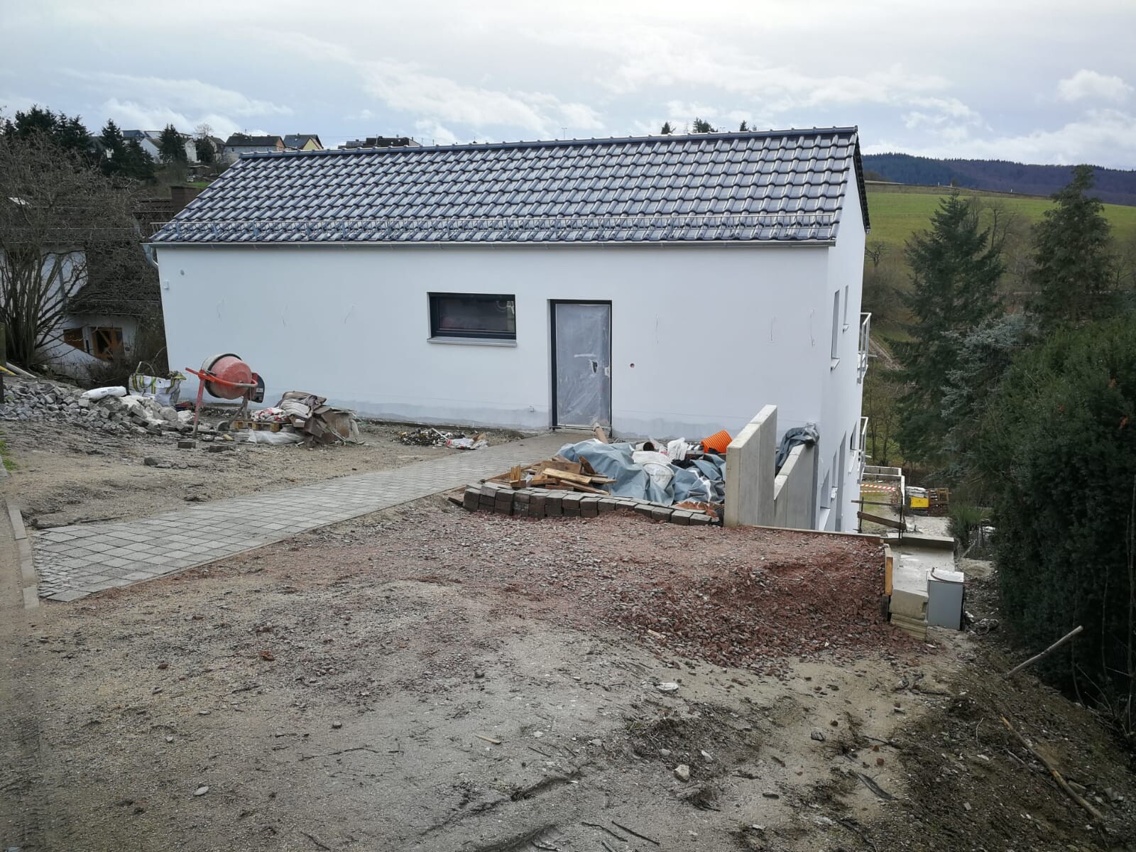White construction site on hillside with roof tiles, piles of building materials and cement mixer on the left.
