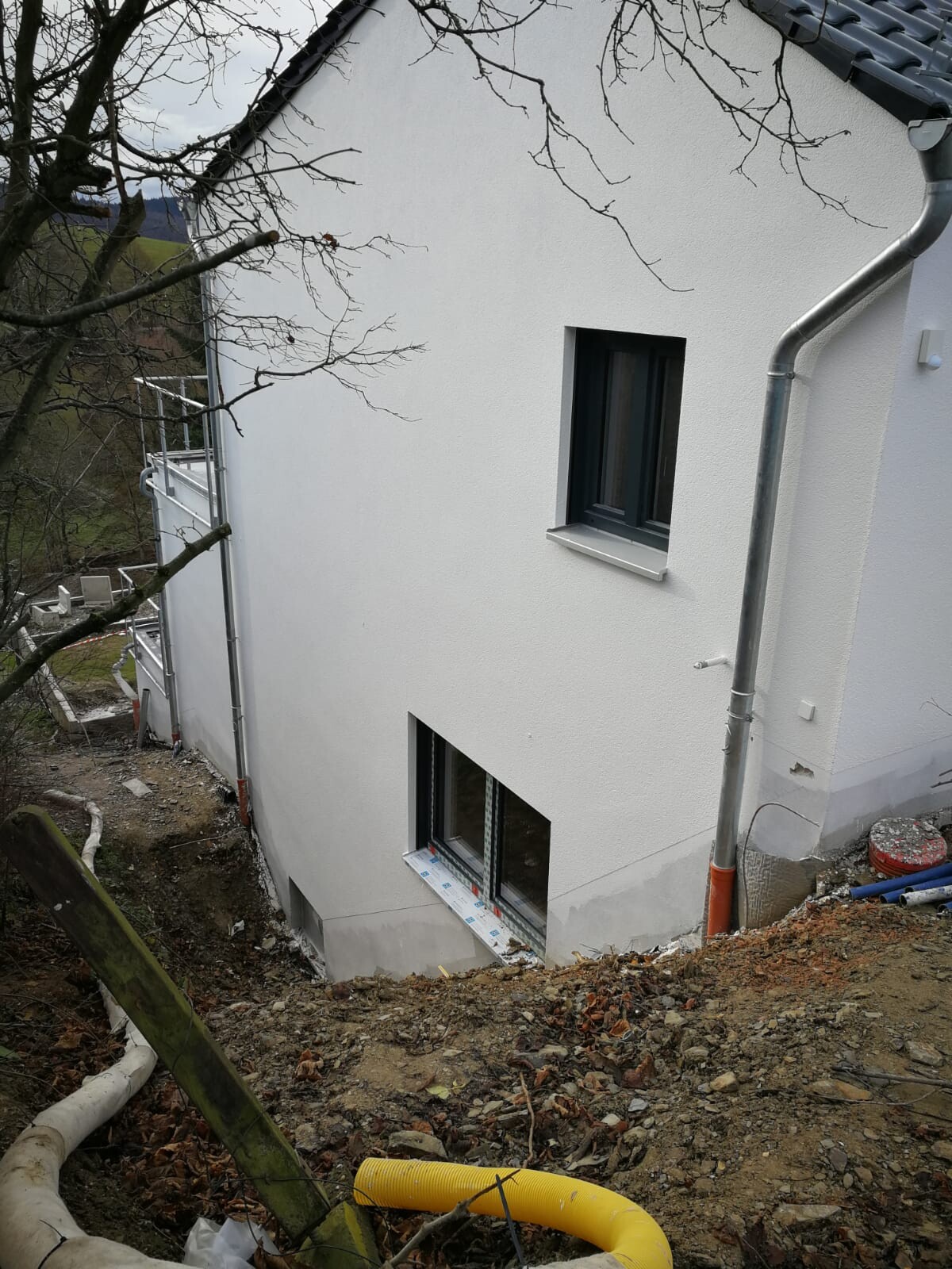 White building facade under construction: two windows (small upper, larger lower), earthworks.