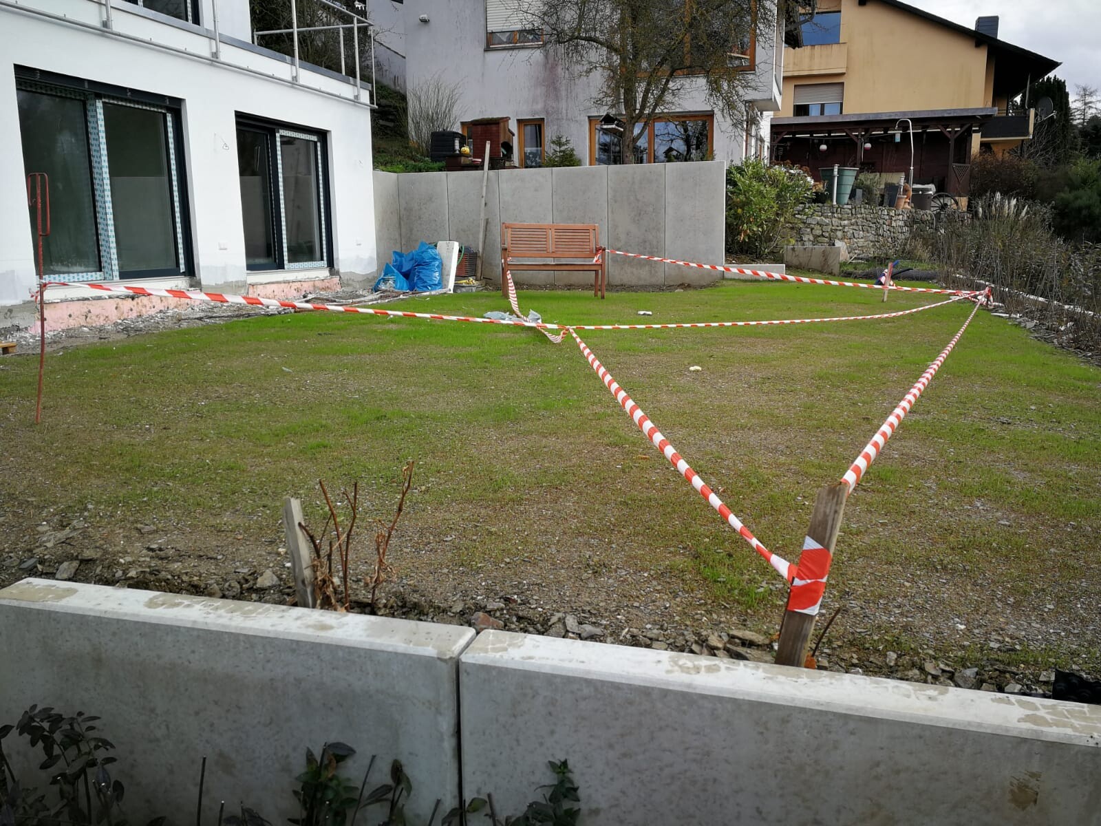Garden behind house with red barrier tape around a building zone; bench and building tools visible.