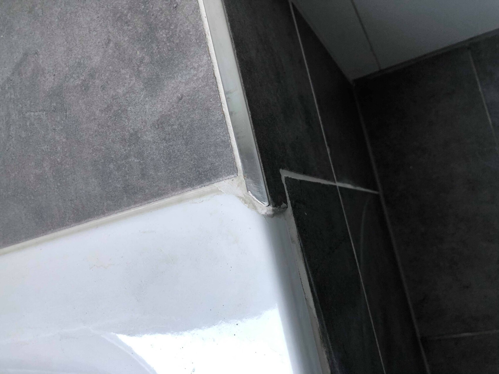 Corner area of a bathtub with gray wall tiles, white edge, and dark floor, metal trim.